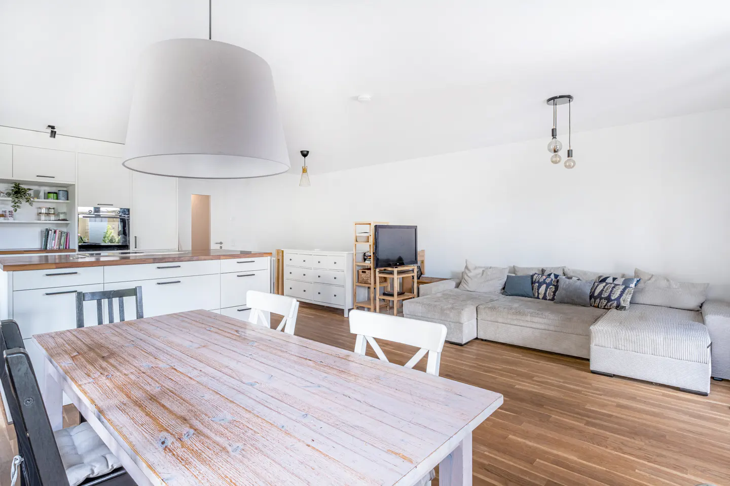 Open-plan living area with a white kitchen, wooden dining table, and a large grey sectional sofa on hardwood floors.