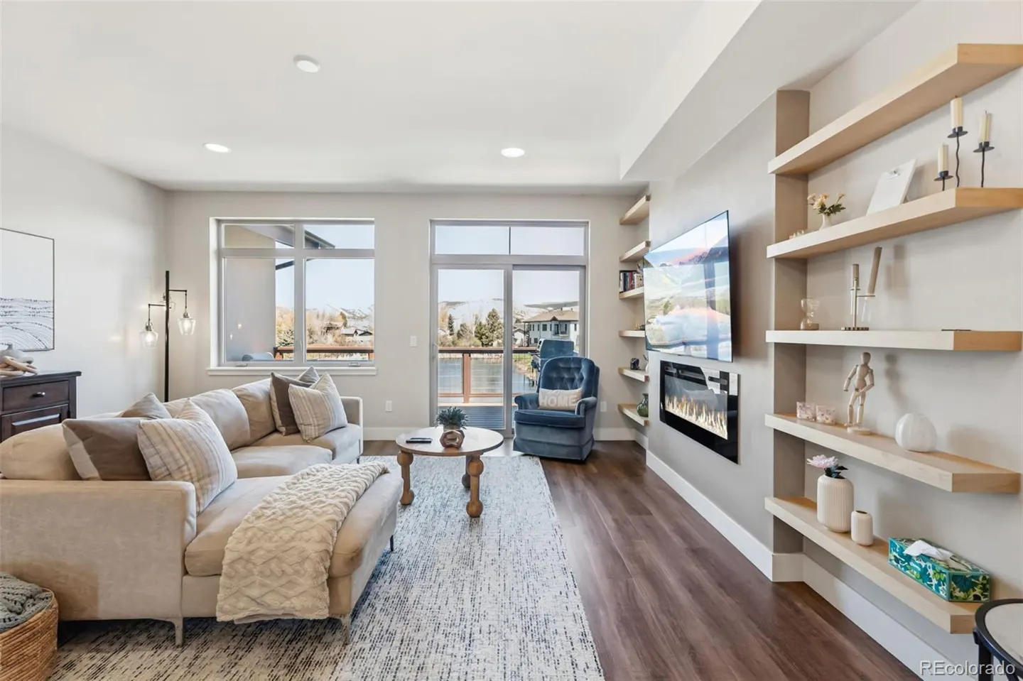 Living room with a beige sofa, blue armchair, and light wood shelves. A TV and fireplace are mounted on the wall. Large windows offer a view of a lake.