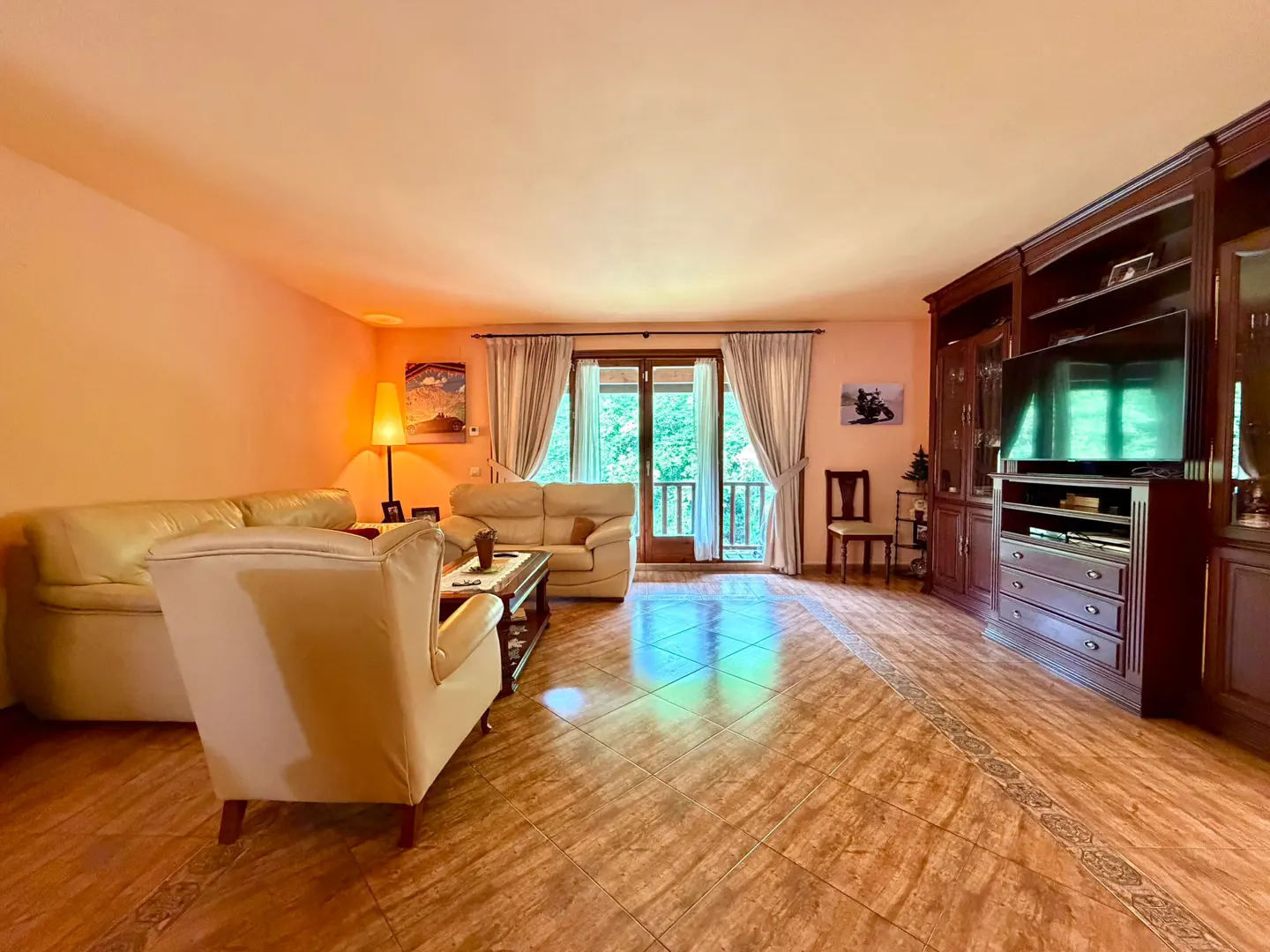 Living room with tile floor, cream sofas, and dark wood built-in shelving with a TV. Balcony doors with curtains lead to greenery.