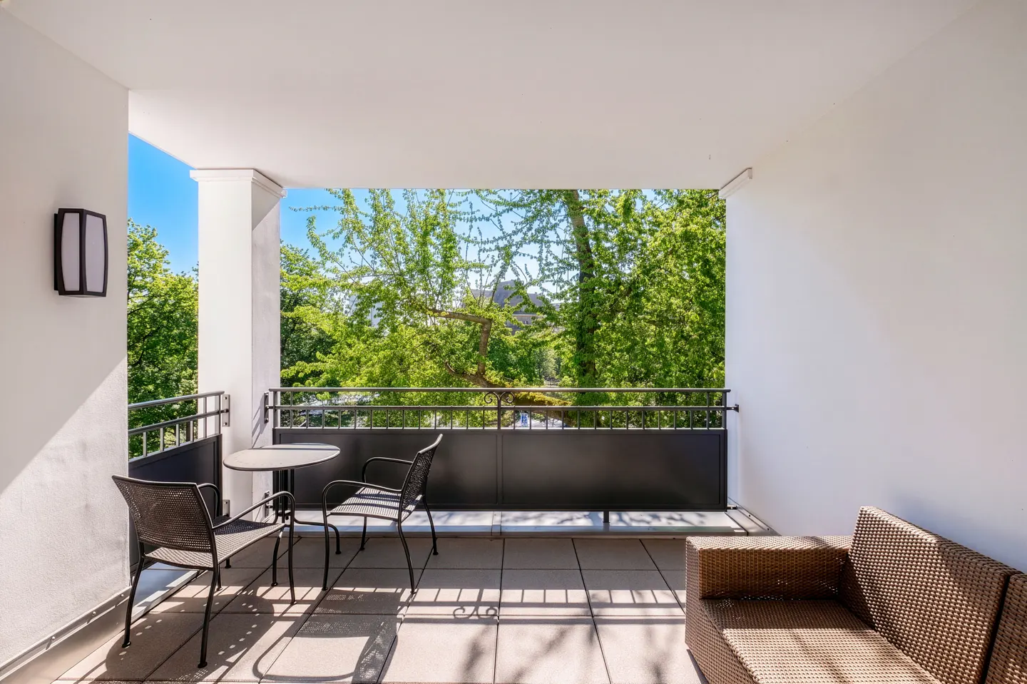 Covered balcony with a small table, two metal chairs, and a wicker sofa overlooking lush green trees under a blue sky.