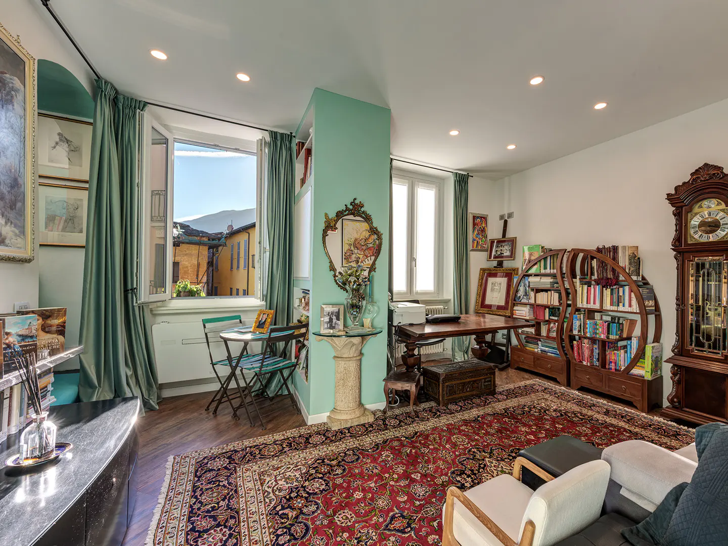 Bright living room with a red patterned rug, a wooden desk, and a grandfather clock. A window with green curtains overlooks a European street.