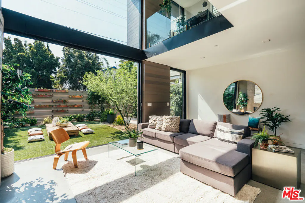 Bright living room with a gray sectional sofa, round mirror, and a view of a green backyard with a wooden table and chairs.