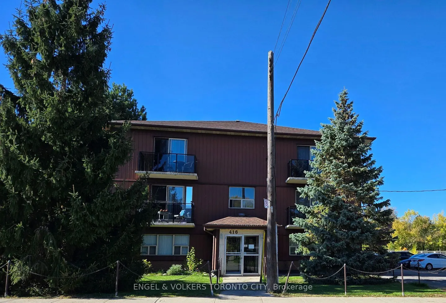 Three-story brown apartment building with balconies, surrounded by large pine trees under a clear blue sky.
