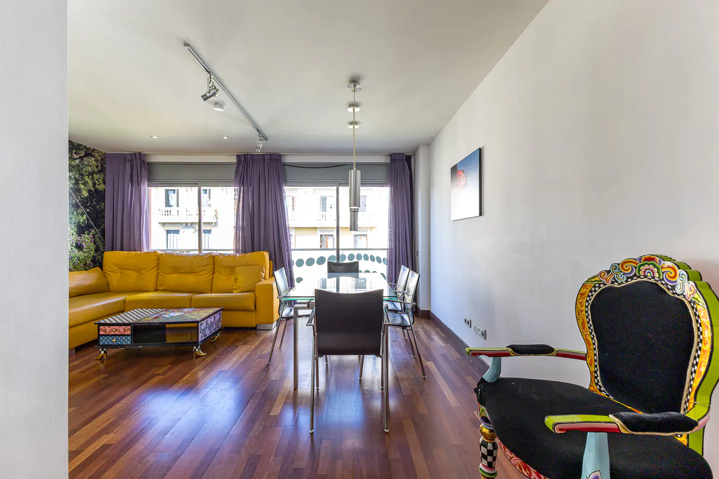 Bright living room with yellow sofa, glass table, and ornate chair. Purple curtains frame the window. Hardwood floors add warmth.