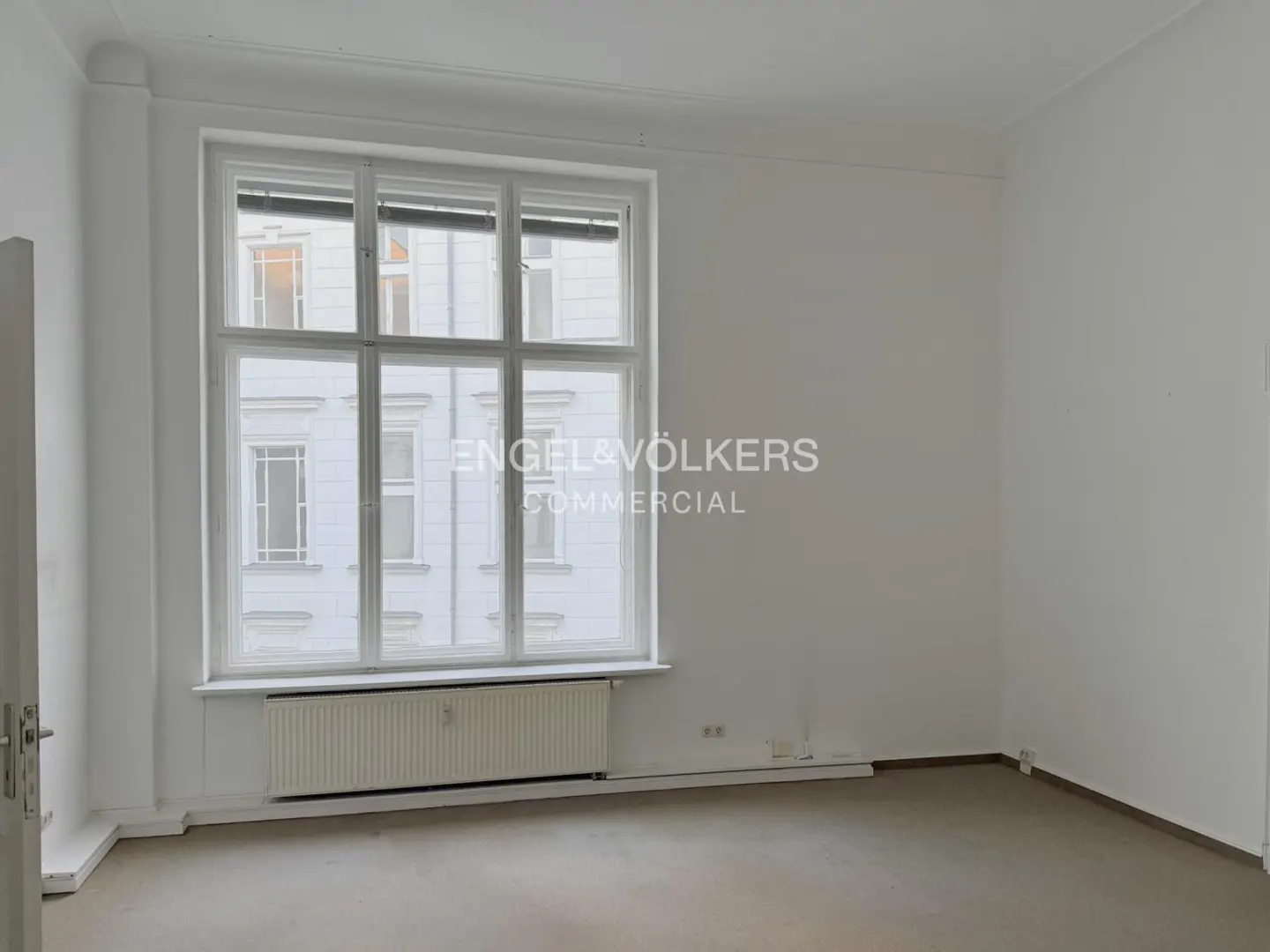 Bright, empty room with white walls, a large window, and a radiator. The Engel & Völkers logo is superimposed on the window.