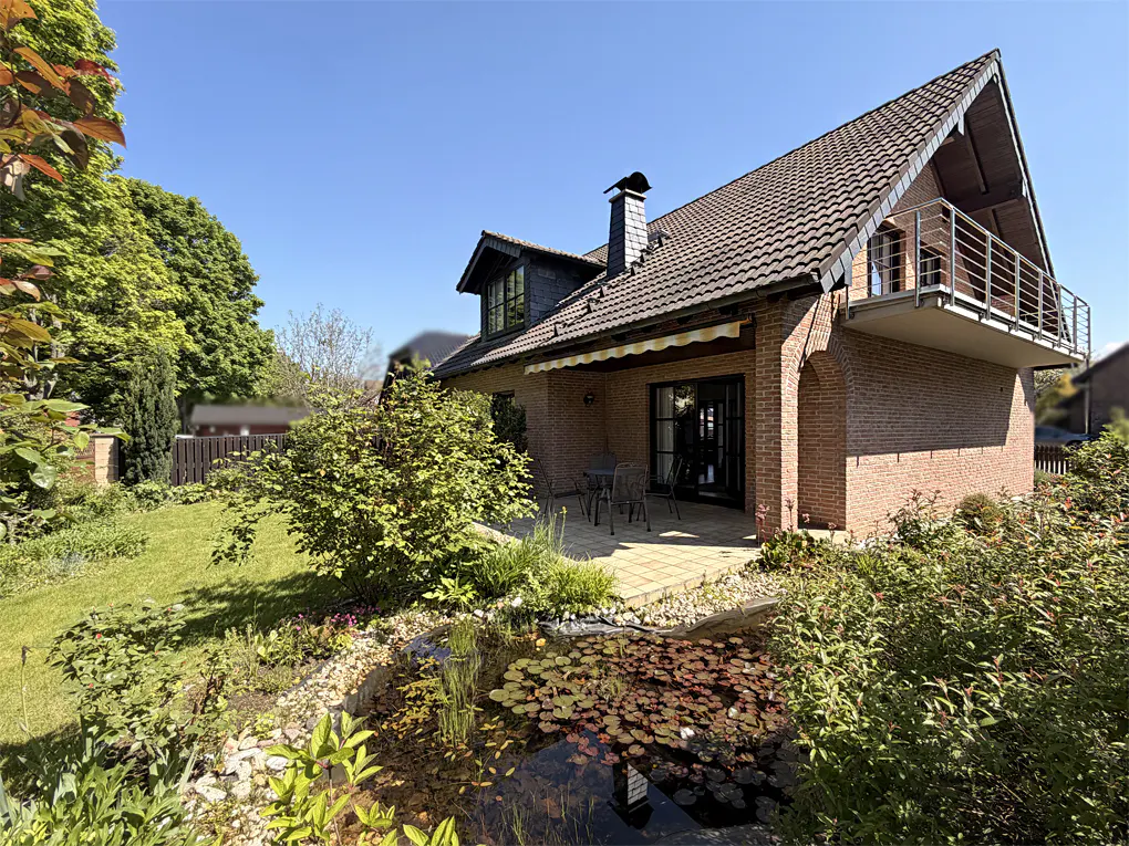 Exterior view of a brick house with a pond, patio, and balcony on a sunny day.