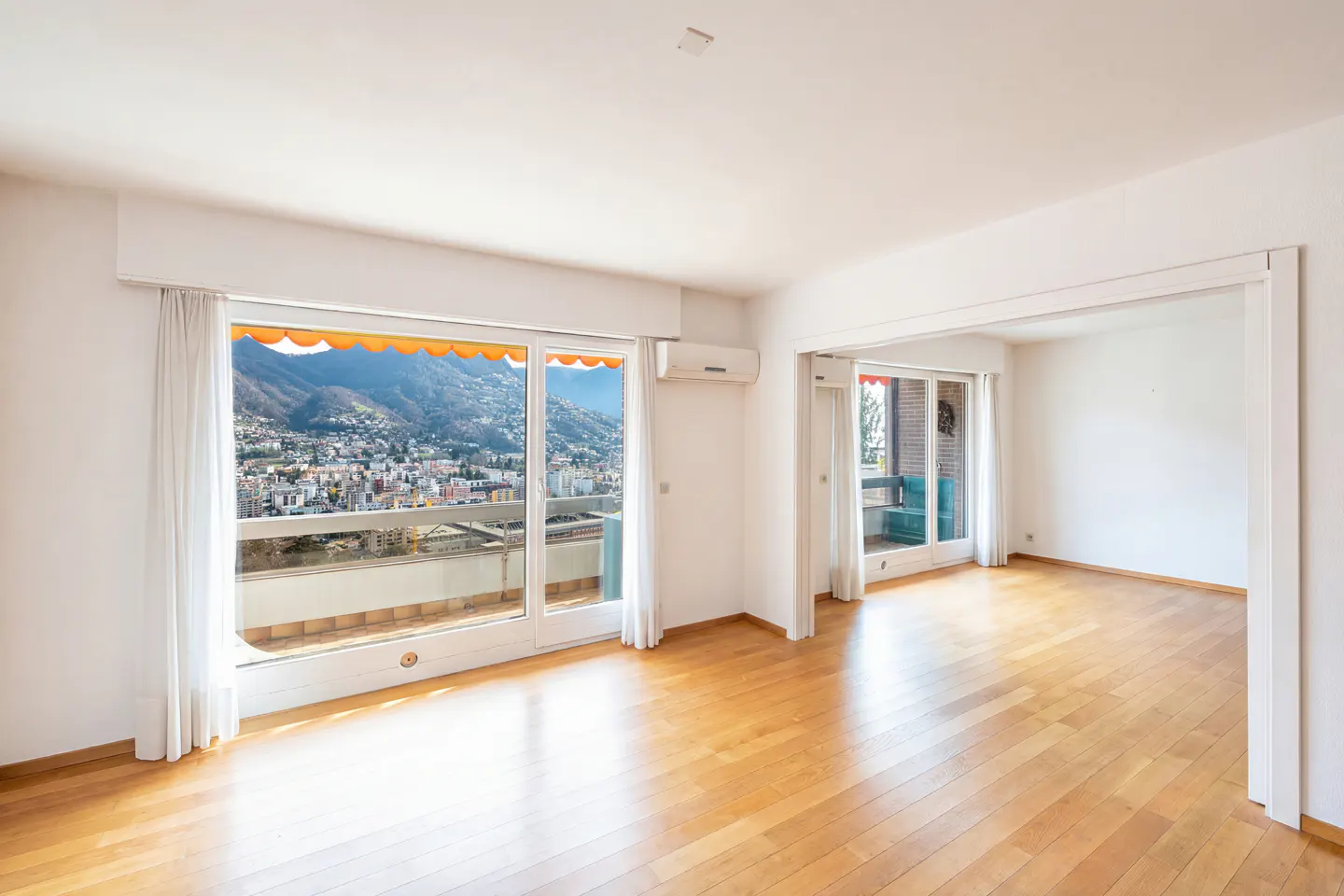 Bright, empty room with wood floors and large windows. A city and mountains are visible through the windows.
