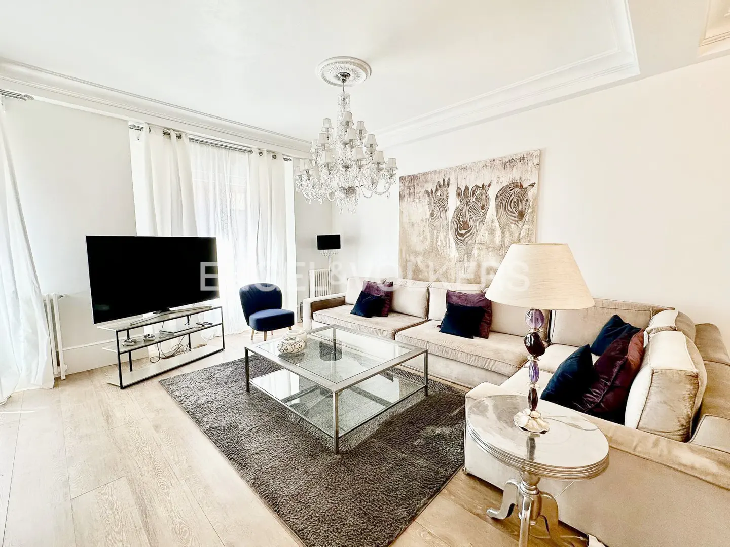Bright living room with a beige sectional sofa, glass coffee table, large zebra art, and a crystal chandelier.
