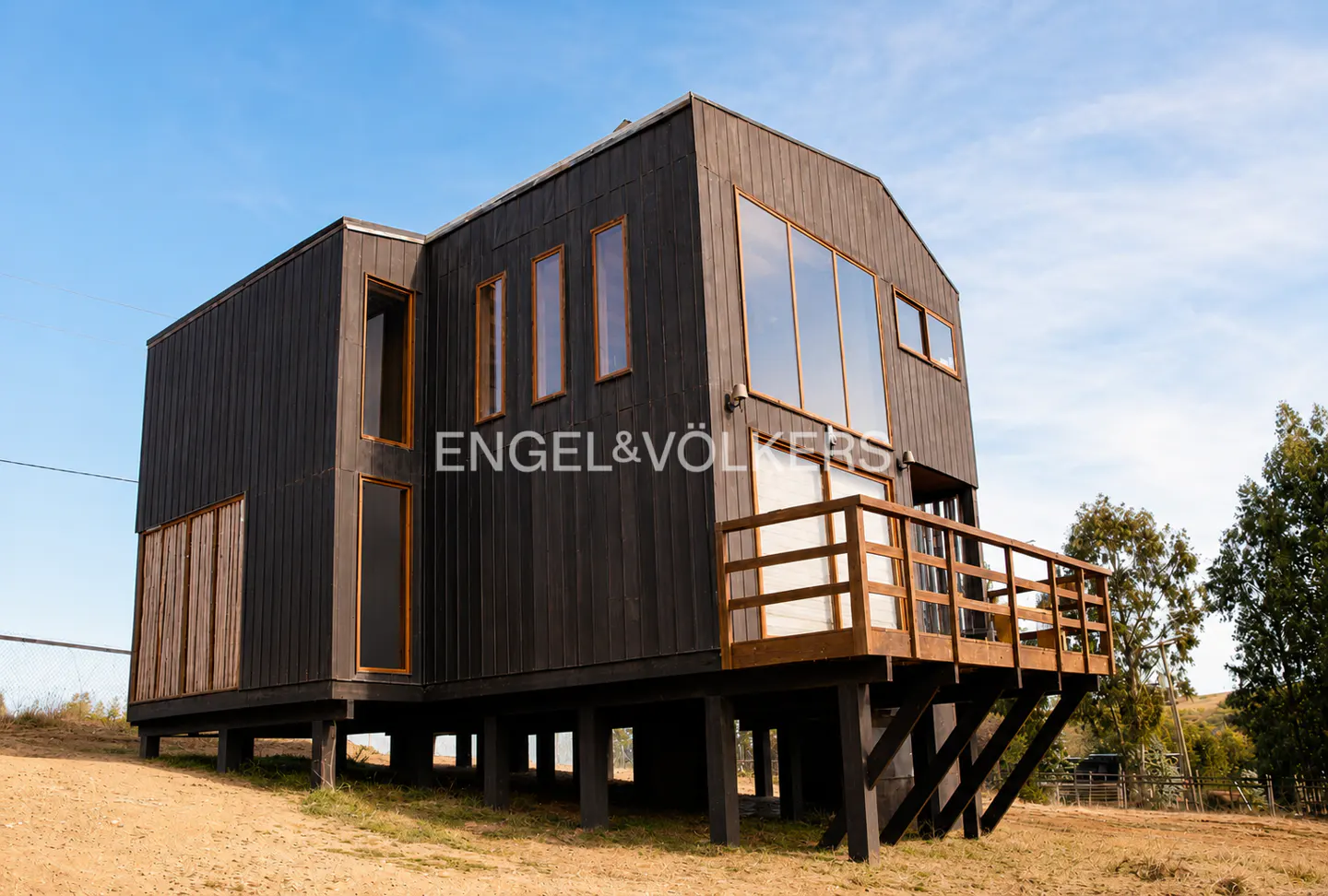 Modern two-story house with dark wood siding, large windows, and a wooden deck, set on stilts in a rural landscape.