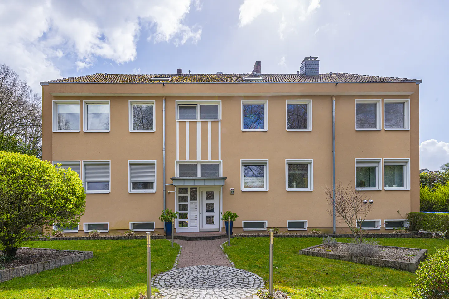 Exterior view of a three-story, tan apartment building with white trim and a brick walkway leading to the front door.