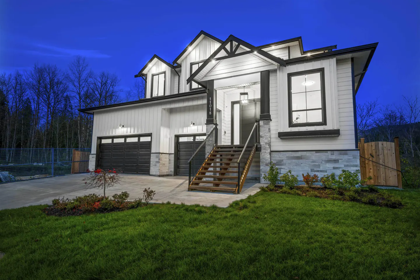 Two-story white house with black trim, a two-car garage, and a wooden staircase leading to the front door. Green lawn and trees surround the house.
