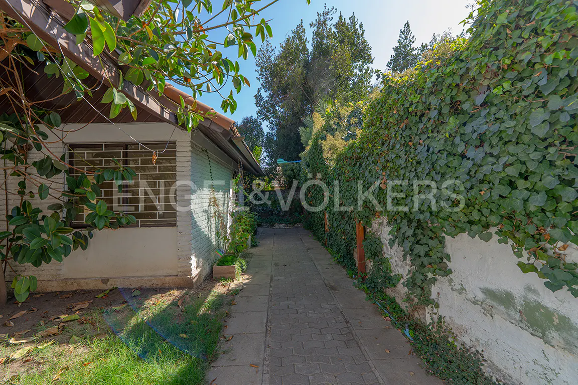A stone path leads between a white brick house and a vine-covered wall under a blue sky. Trees are visible in the background.
