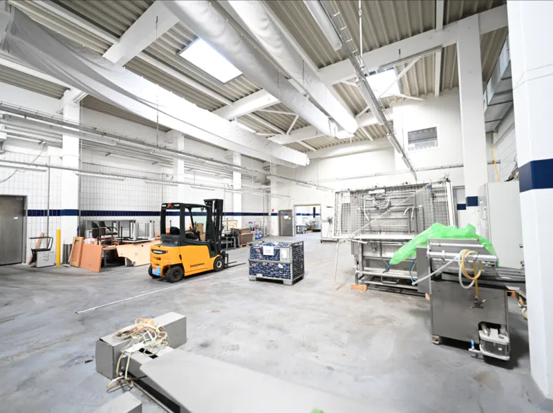 Bright, spacious warehouse interior with a yellow forklift, machinery, and white walls. Skylights illuminate the scene.