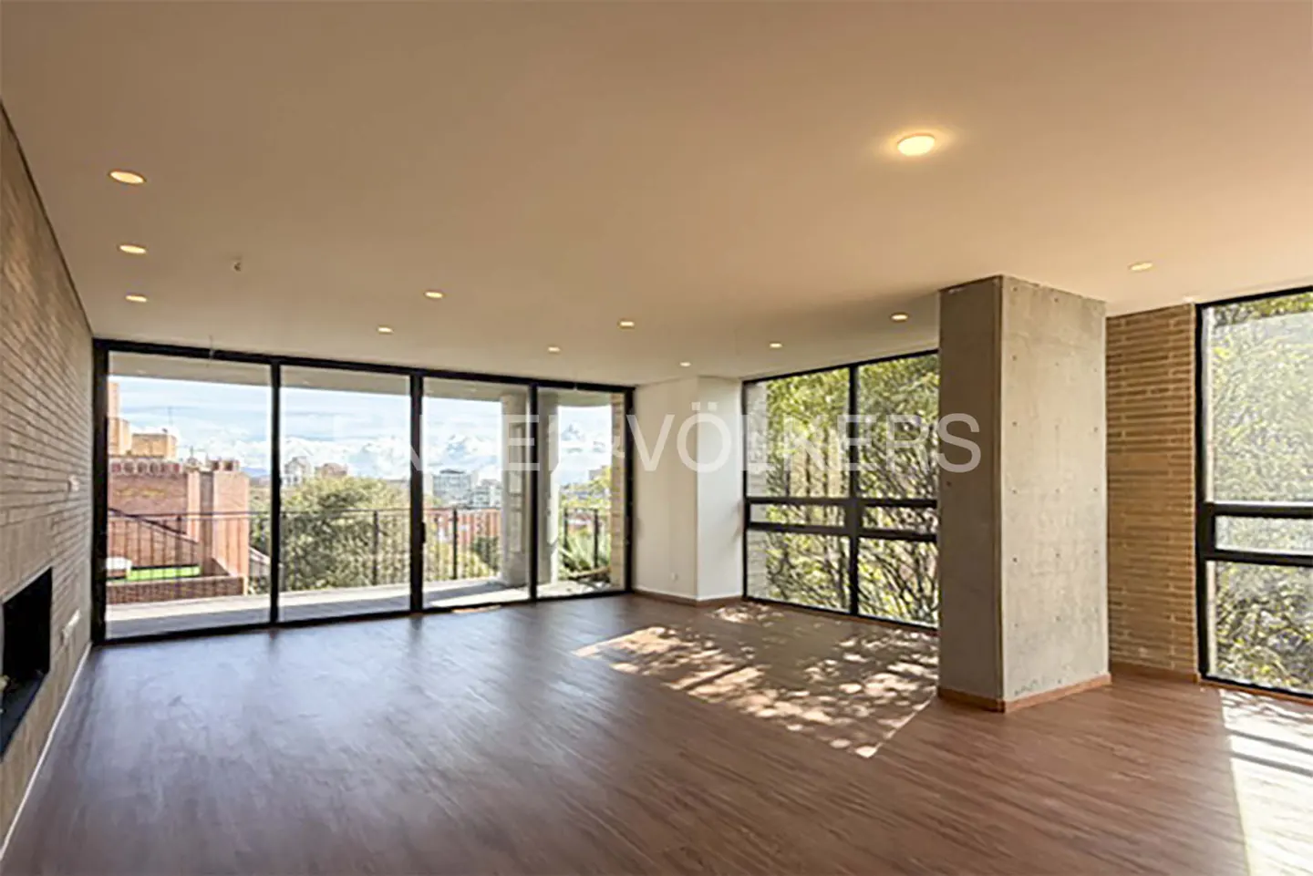 Bright, empty living room with hardwood floors, brick walls, and large windows with city views.