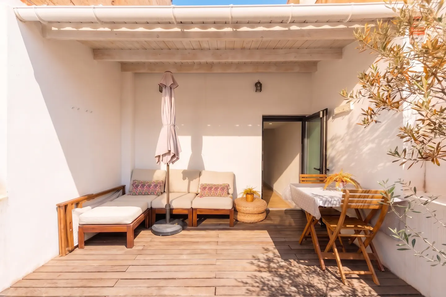Outdoor patio with wooden deck, white walls, beige lounge seating, and a table with chairs. An umbrella stands next to the lounge.