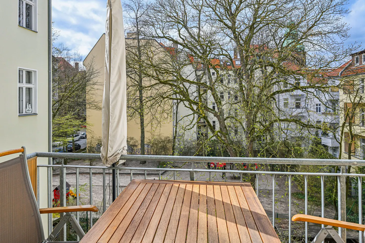Balcony view with a wooden table and chairs, overlooking a courtyard with trees and buildings on a sunny day.