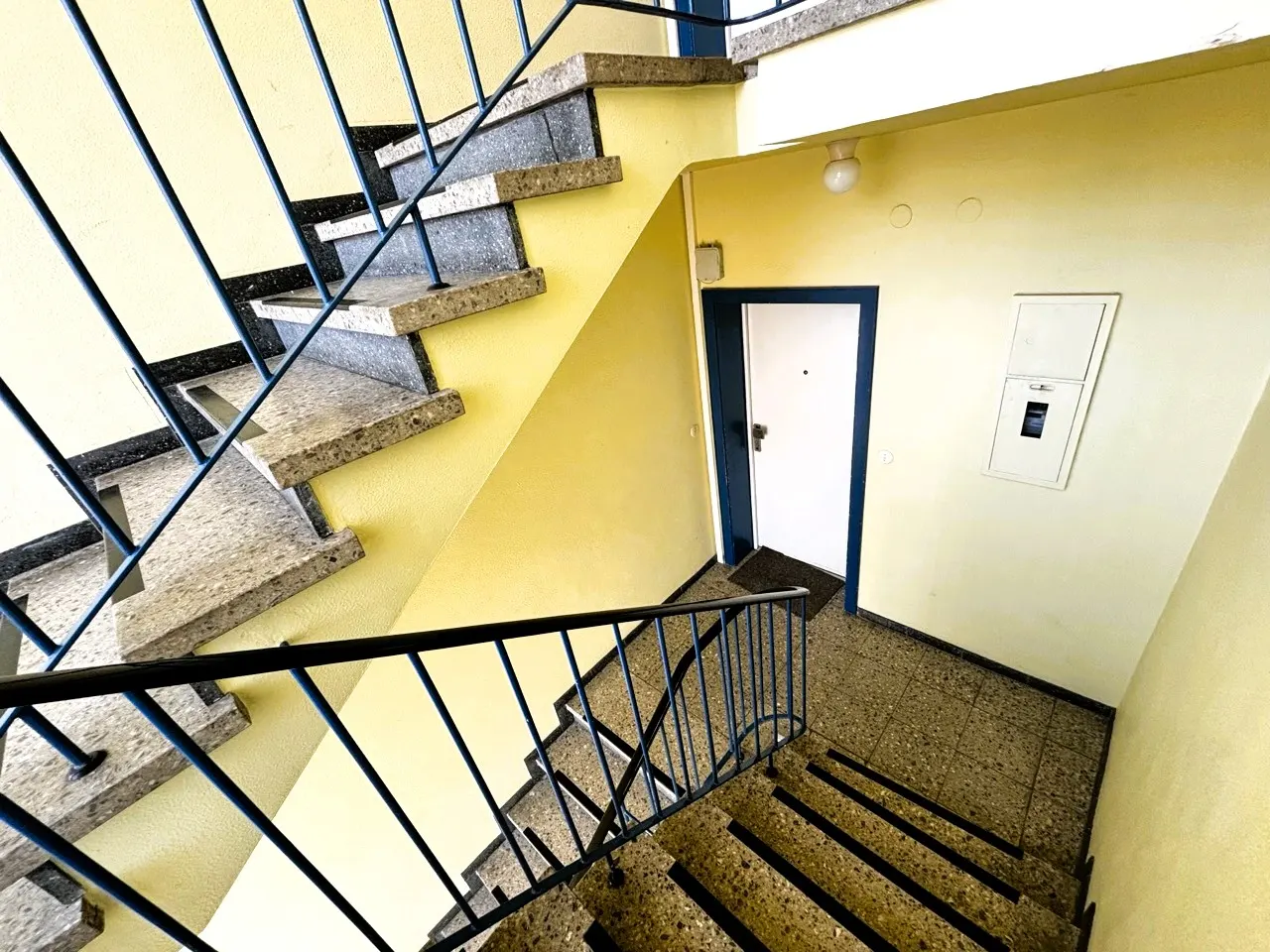 Stairwell with granite steps, blue railings, and yellow walls. A white door with a blue frame is visible.