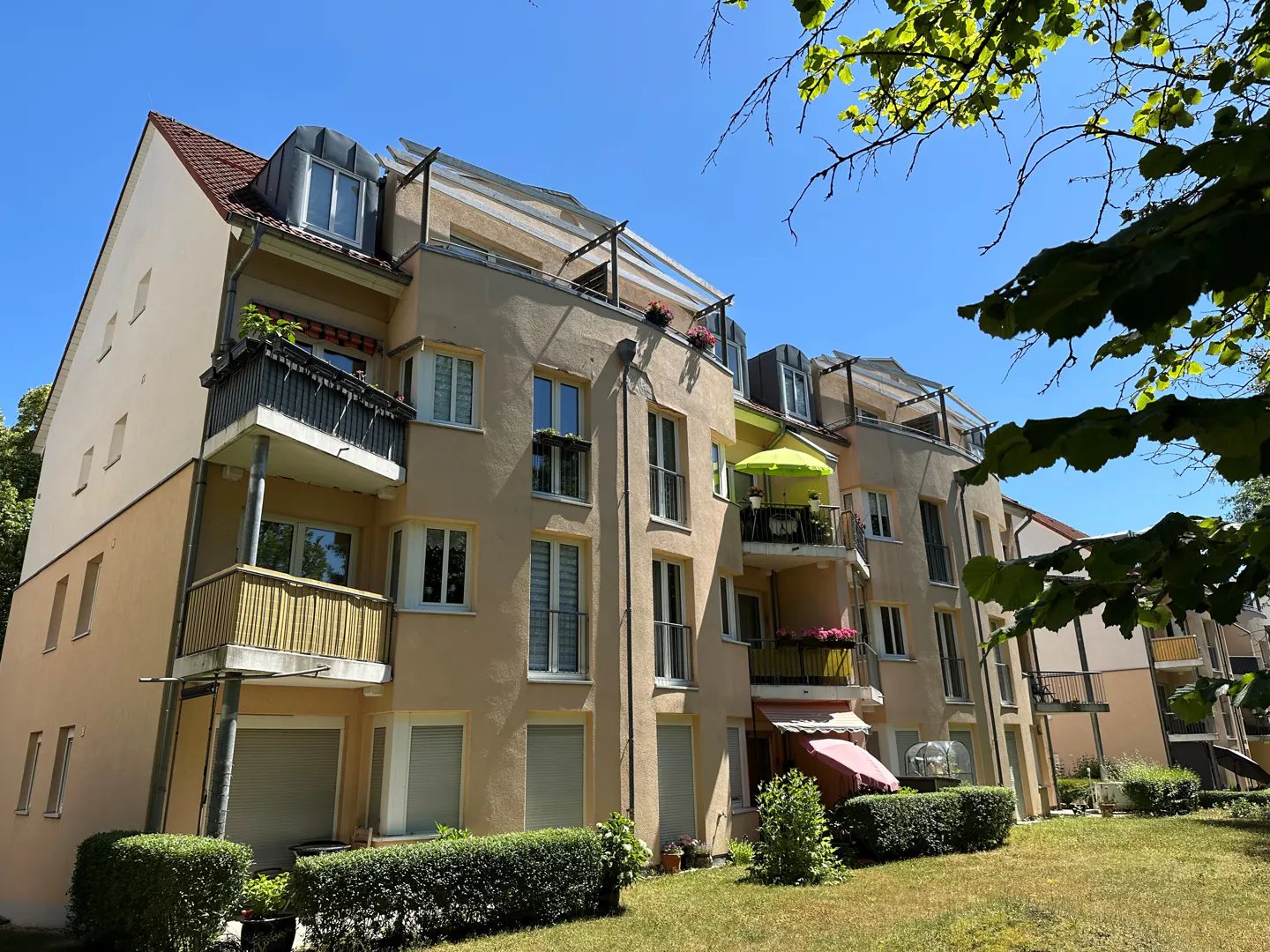Exterior view of a multi-story apartment building with balconies, tan walls, and green lawn under a blue sky.