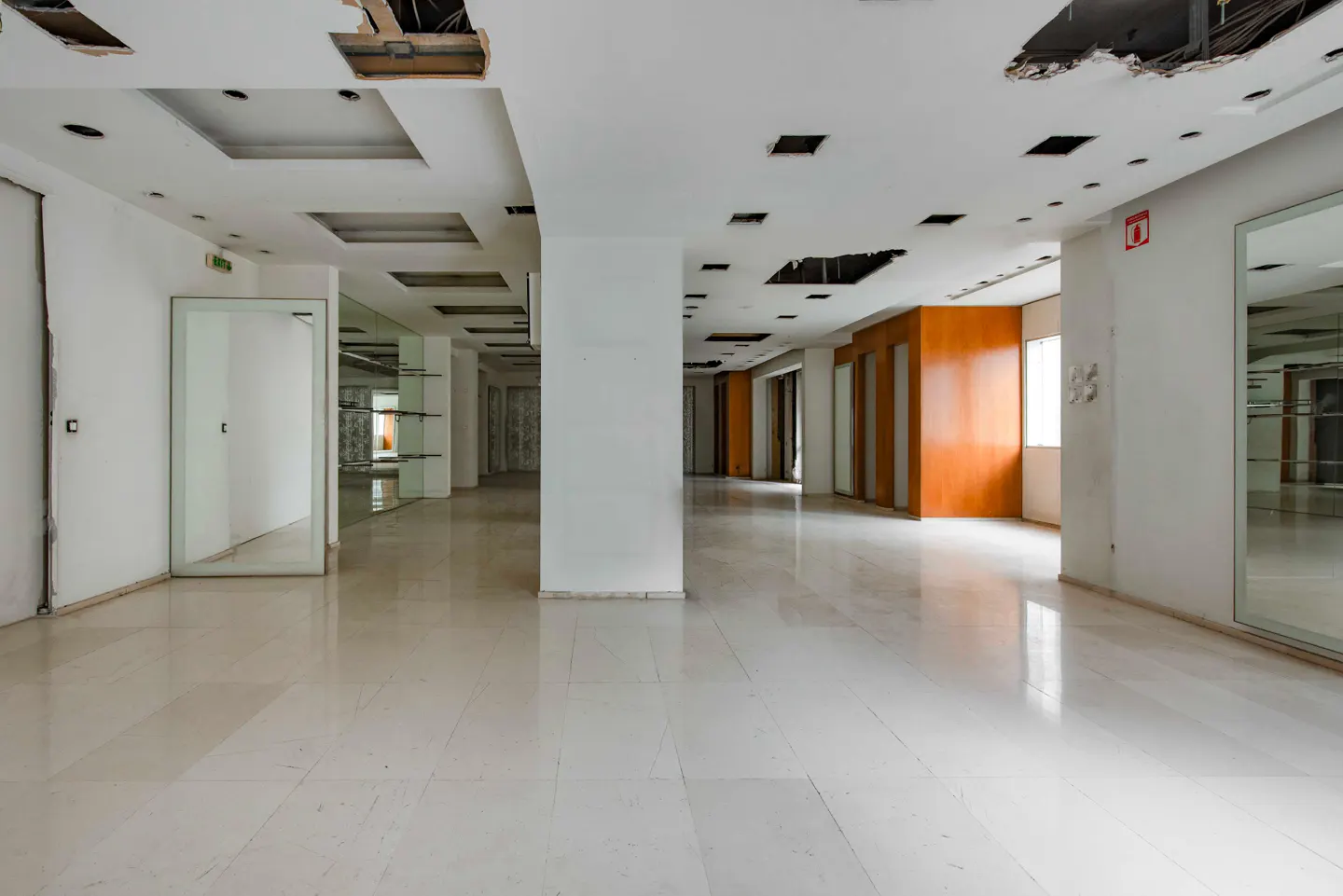 Empty commercial space with white walls, marble floors, and exposed ceiling. Orange accent walls and glass doors line the hallway.