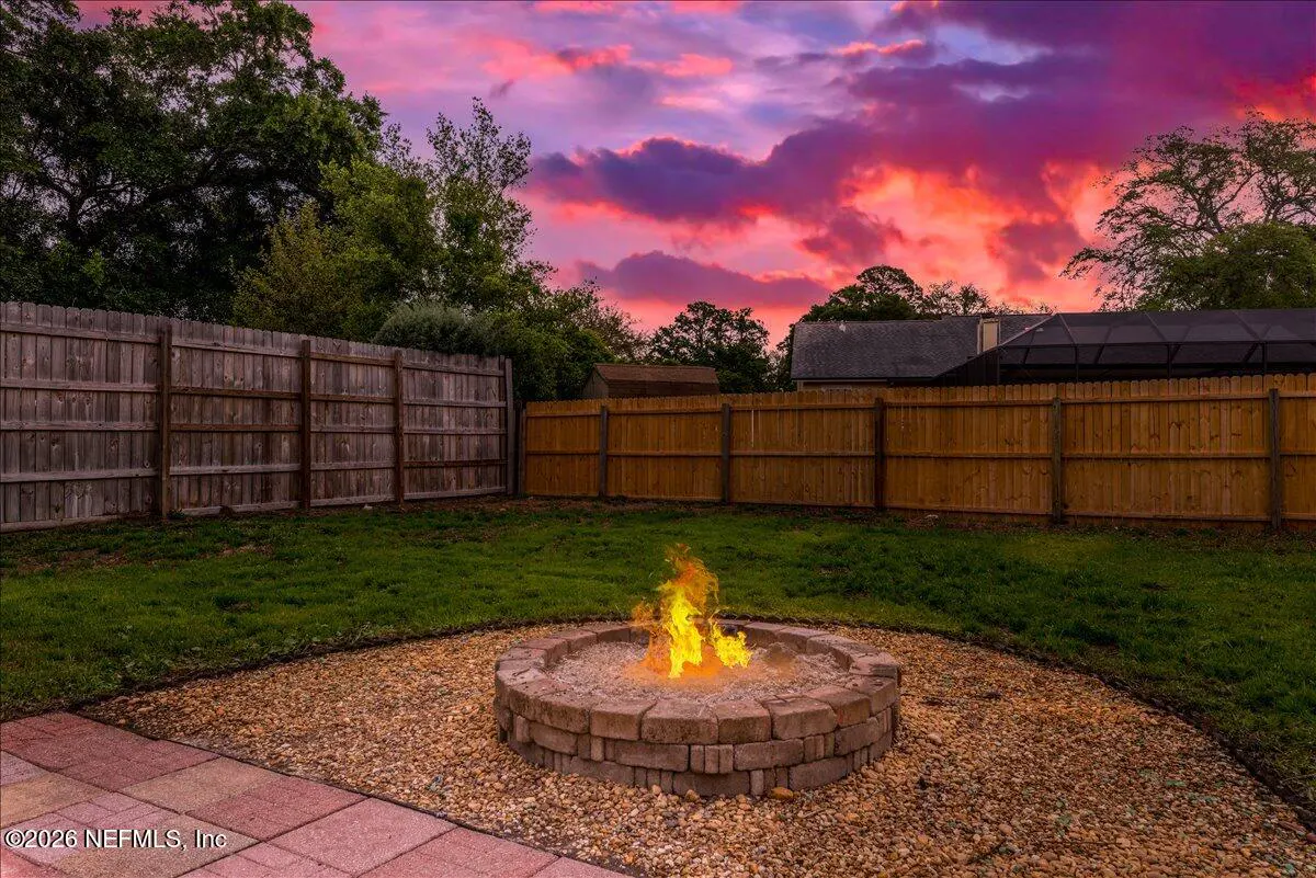 Backyard with a stone fire pit ablaze, surrounded by grass and a wooden fence under a vibrant pink and orange sunset.