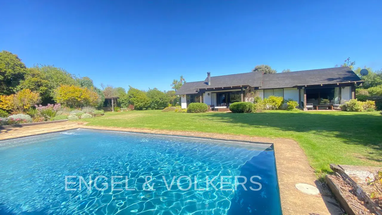 A backyard view of a white house with a pool, green lawn, trees, and a clear blue sky.