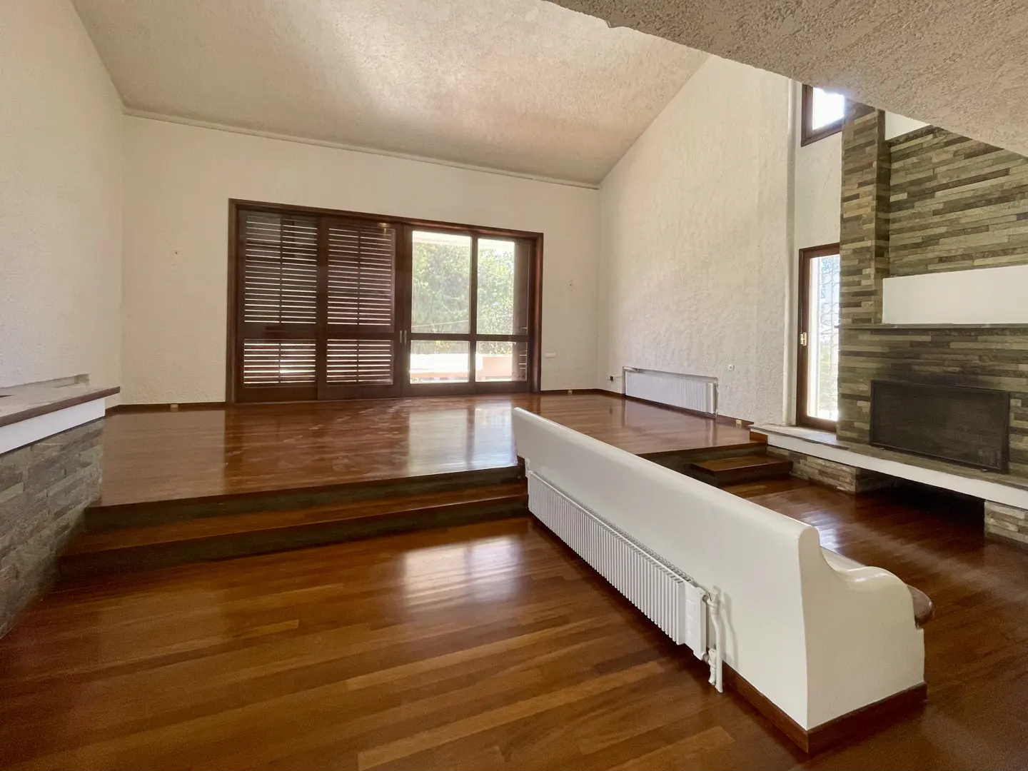 A spacious living room with hardwood floors, a stone fireplace, and a sliding glass door. The room has white walls and a high ceiling.