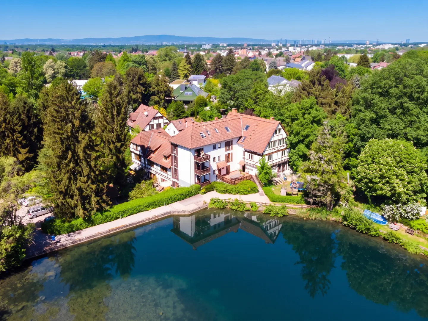 Aerial view of a white building with a red tile roof, surrounded by green trees, next to a blue lake.