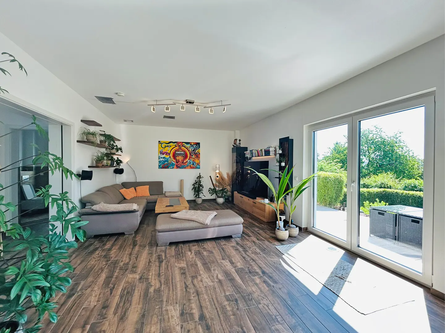 Bright living room with wood floors, a gray sectional sofa, and a colorful painting on the white wall. Large windows overlook a green garden.