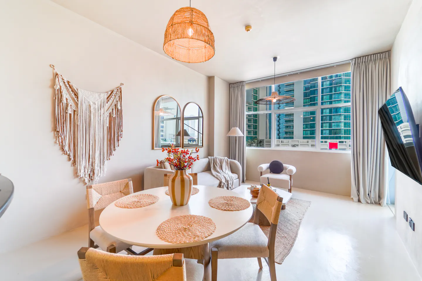 Bright living room with white walls, a round table with woven placemats, and a large window with city views.
