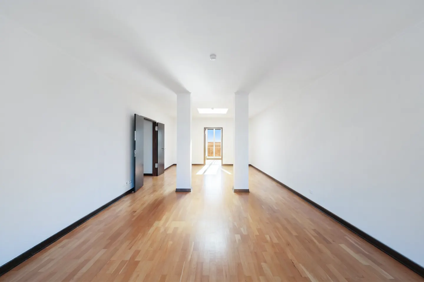 Bright, empty room with hardwood floors, white walls, and dark doors. Columns lead to a window with a view of blue sky.