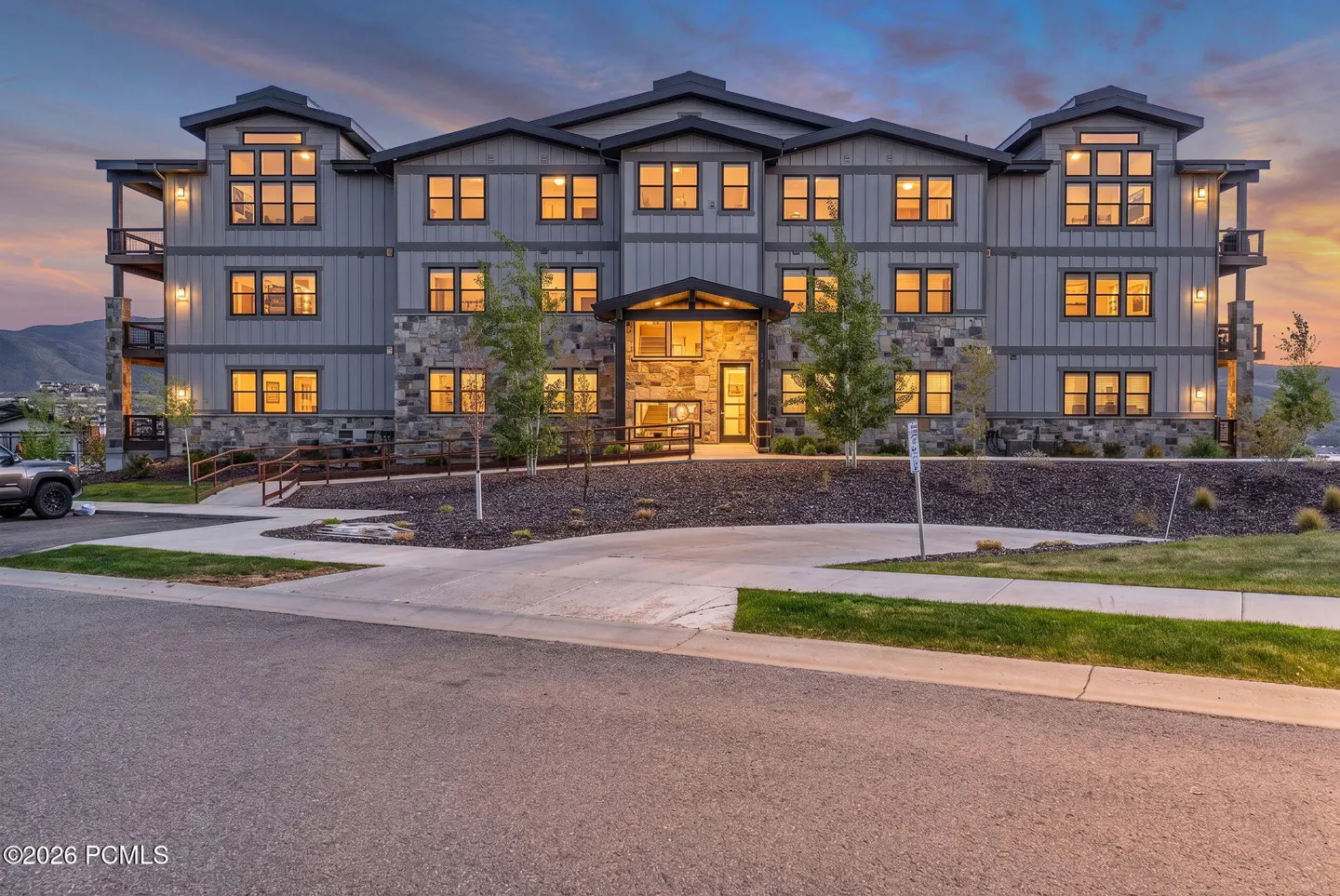 Three-story gray apartment building with stone accents and warmly lit windows at dusk.