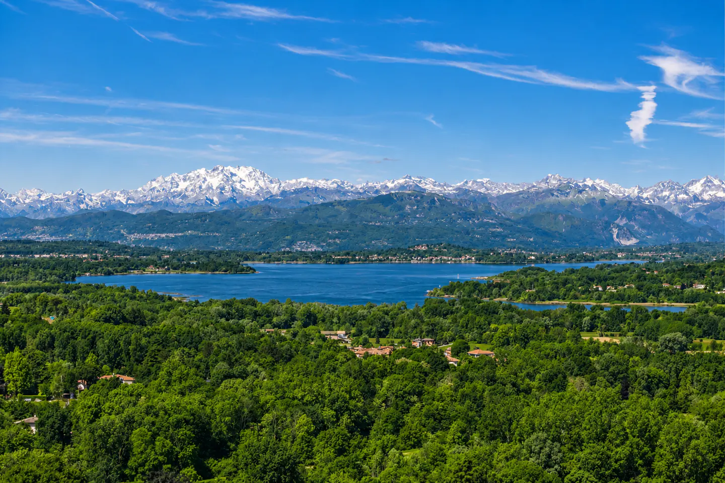 Panoramic view of a blue lake surrounded by lush green forests with snow-capped mountains under a clear blue sky.