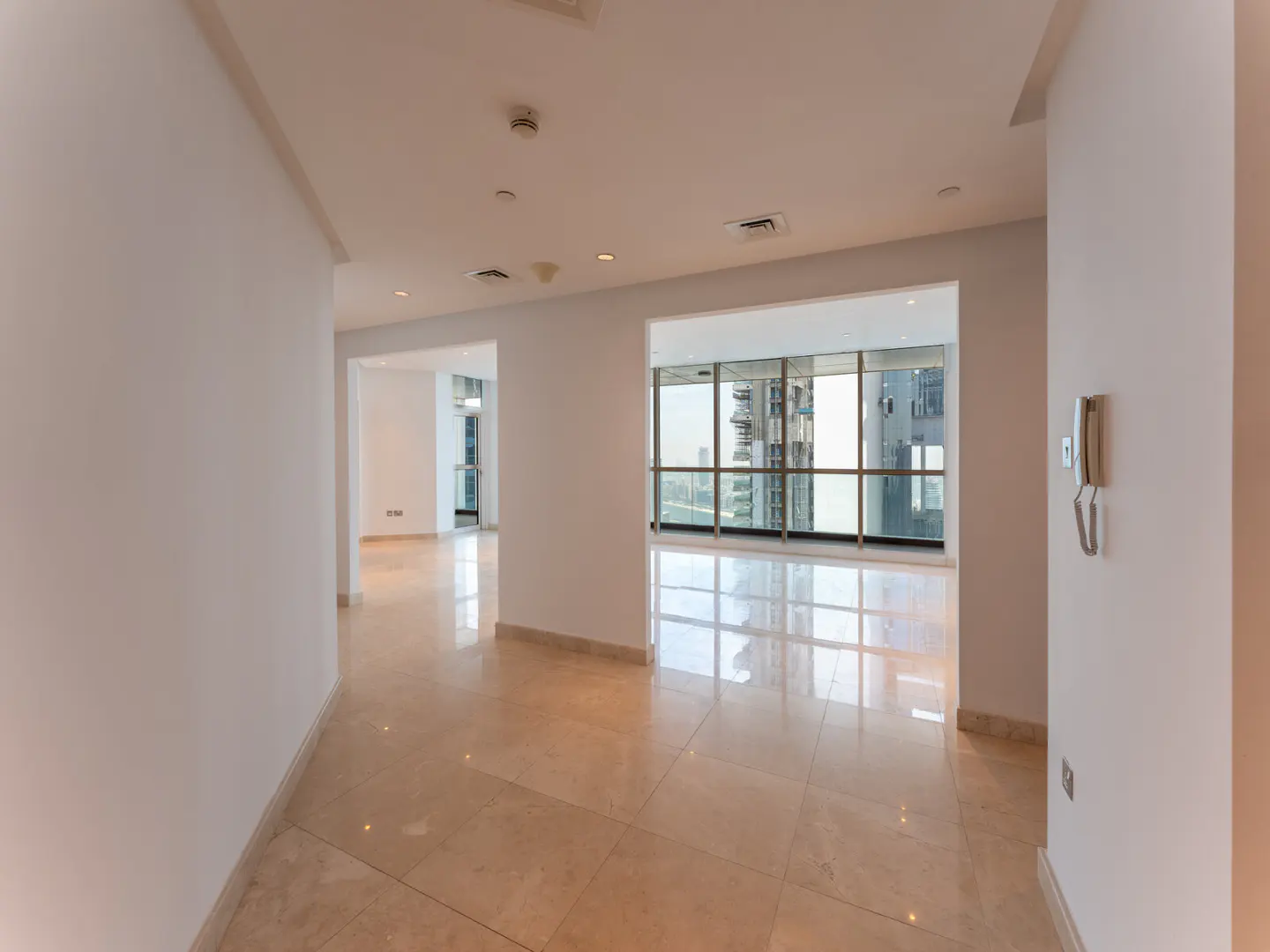 Empty apartment with beige tile floors, white walls, and large windows with city views. A wall-mounted phone is visible.
