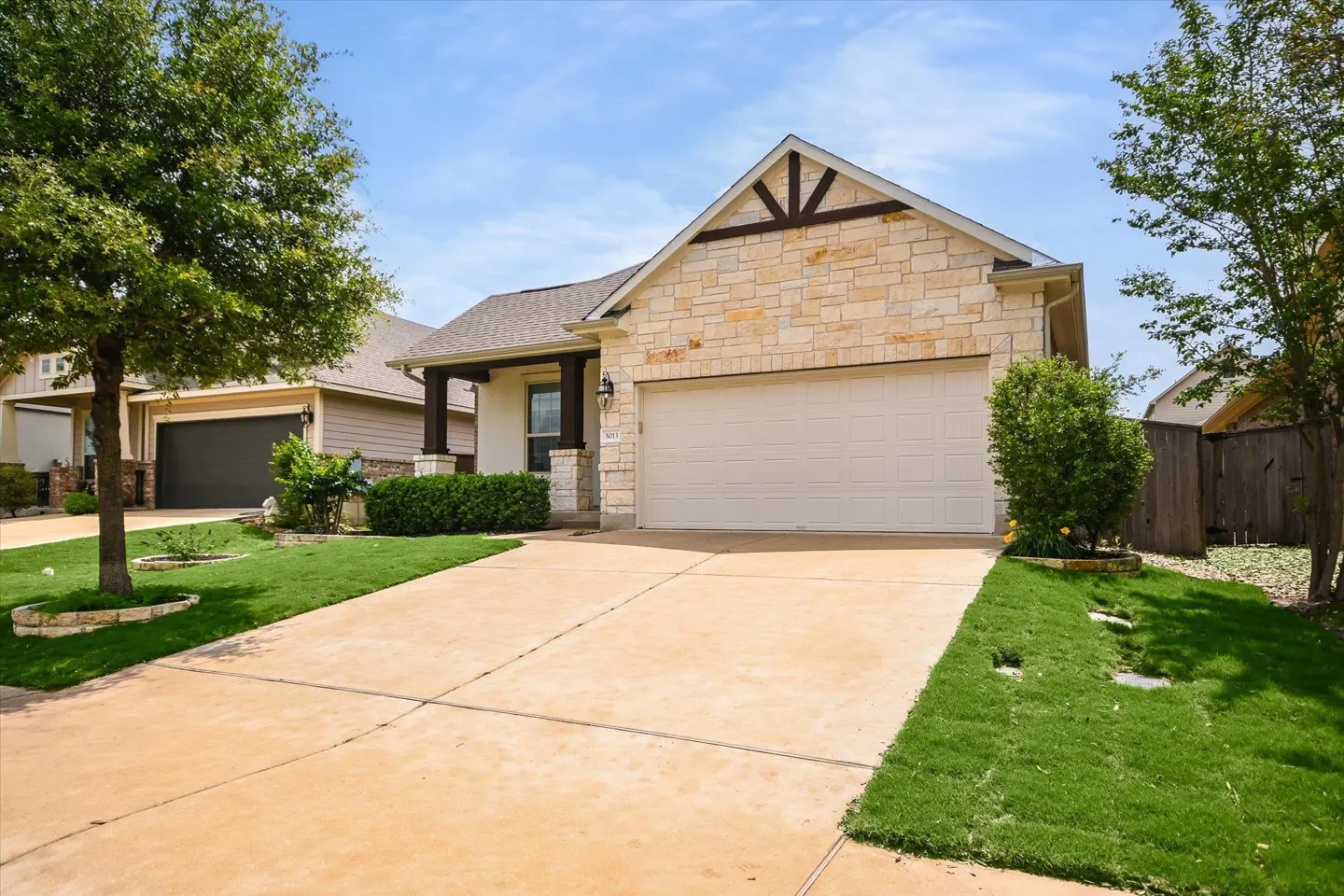 A single-story house with a stone facade, a beige garage door, and a concrete driveway. Green grass and trees surround the house.