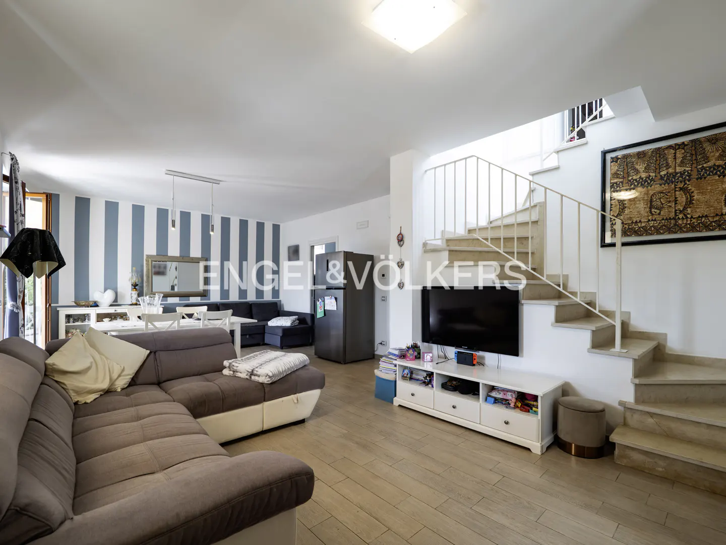 Open-concept living room with a gray sectional sofa, dining table, and staircase. The walls are white with blue stripes. A TV and fridge are visible.