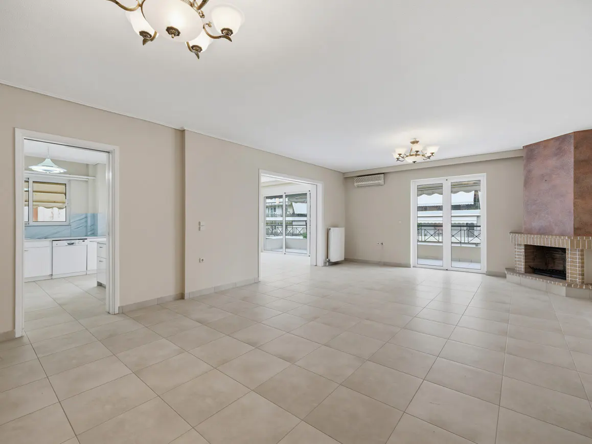 A spacious, empty living room with beige tile flooring, a fireplace, and access to a balcony. A kitchen is visible through a doorway.