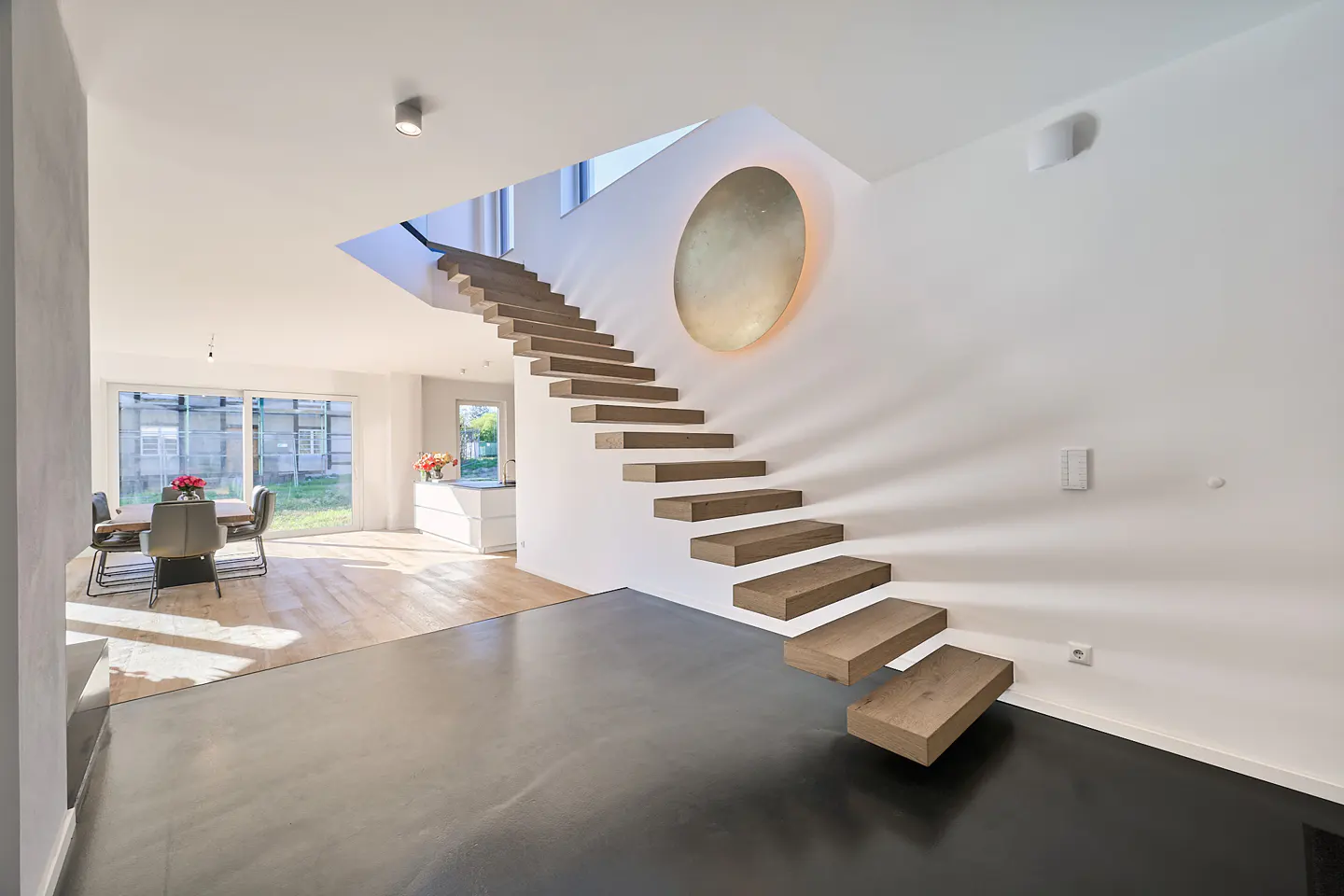 A modern home interior with floating wooden stairs against a white wall, illuminated by a circular light fixture. A dining area is visible in the background.