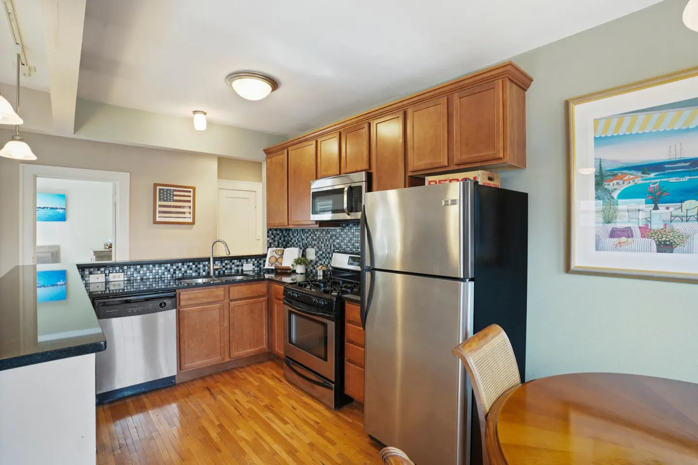 A kitchen with wood cabinets, stainless steel appliances, and a mosaic tile backsplash. A wood table and chair are in the foreground.
