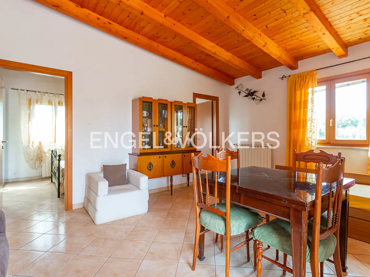 A bright dining room with a wooden ceiling, table, chairs, and a cabinet. A white armchair sits near an open doorway.