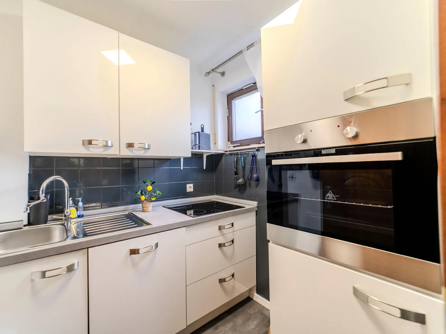 A modern kitchen with white cabinets, stainless steel sink, black backsplash, and built-in oven.