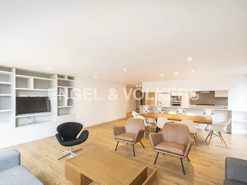 Bright, open-plan living space with wood floors, brown leather chairs, and a long wooden table with white chairs. A black chair sits near a white bookshelf with a TV.