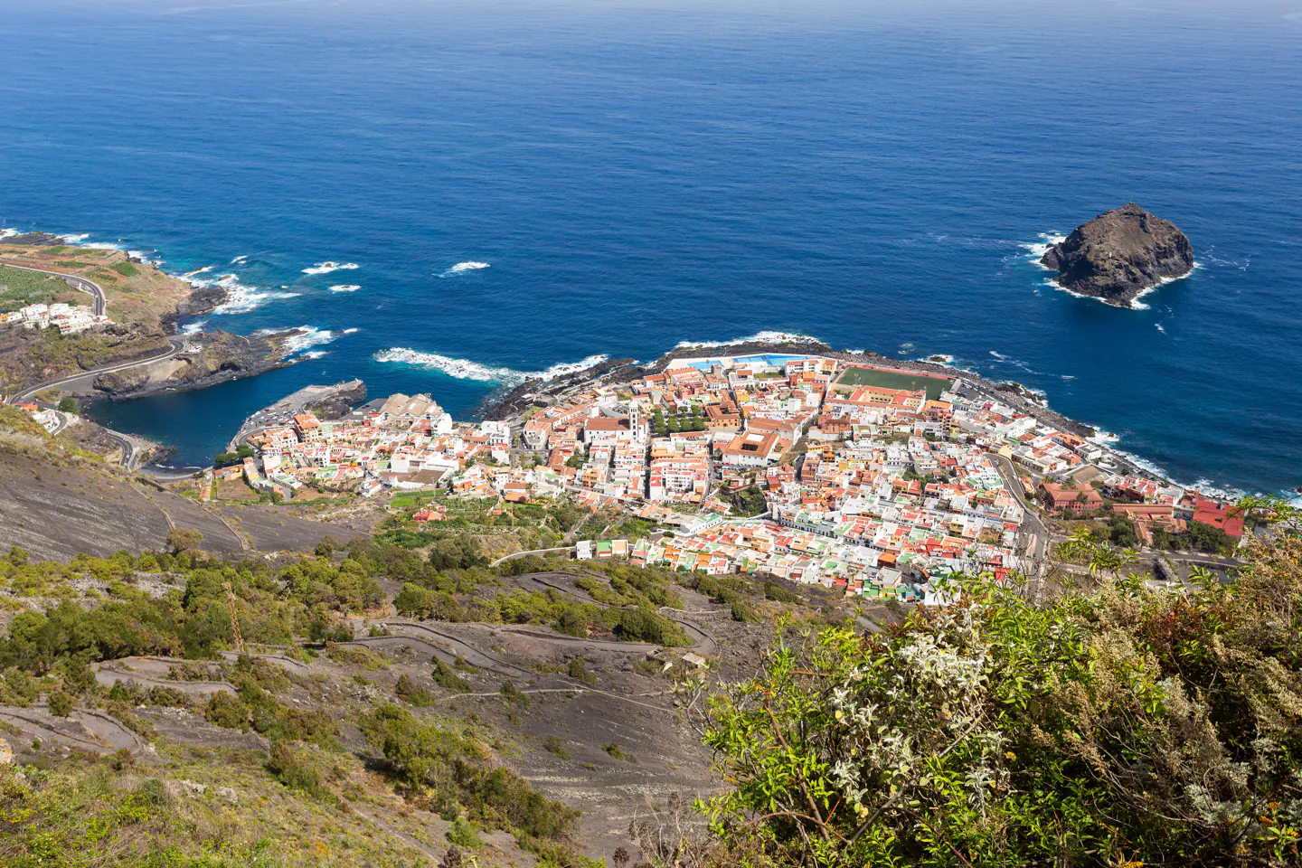 Aerial view of Garachico, Tenerife. White buildings with red roofs line the coast, with blue ocean and a small island in the distance.