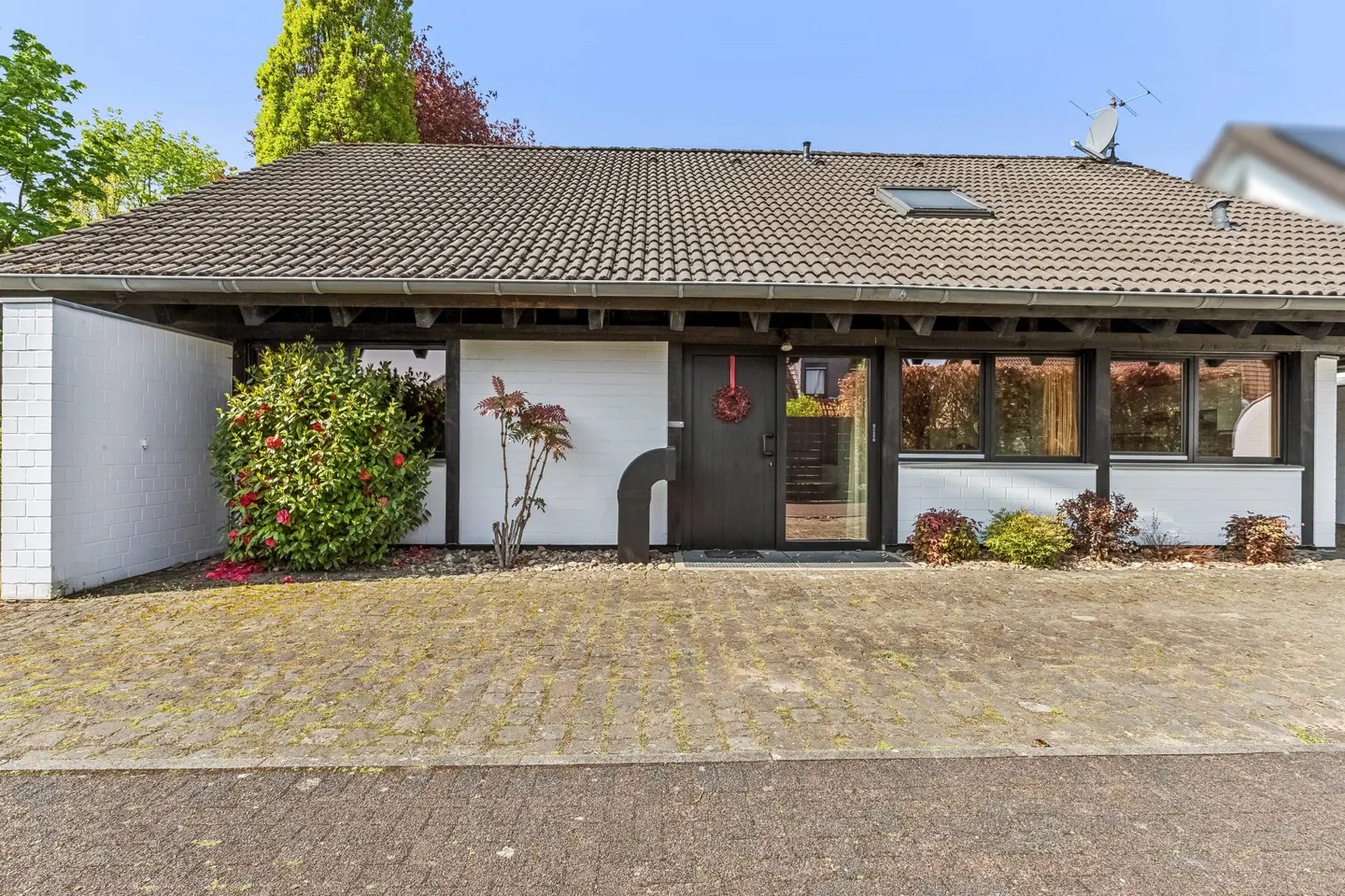 Front exterior of a one-story home with a gray tiled roof, white walls, and black trim. A wreath hangs on the dark front door. A brick driveway leads to the entrance.
