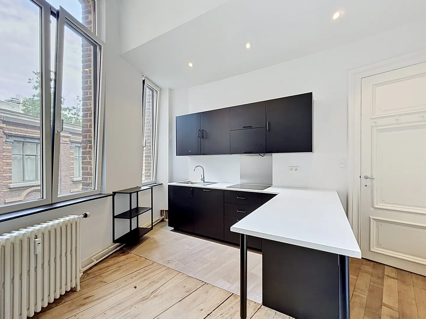 Modern kitchen with black cabinets, white countertops, wooden floors, and large windows overlooking a brick building.