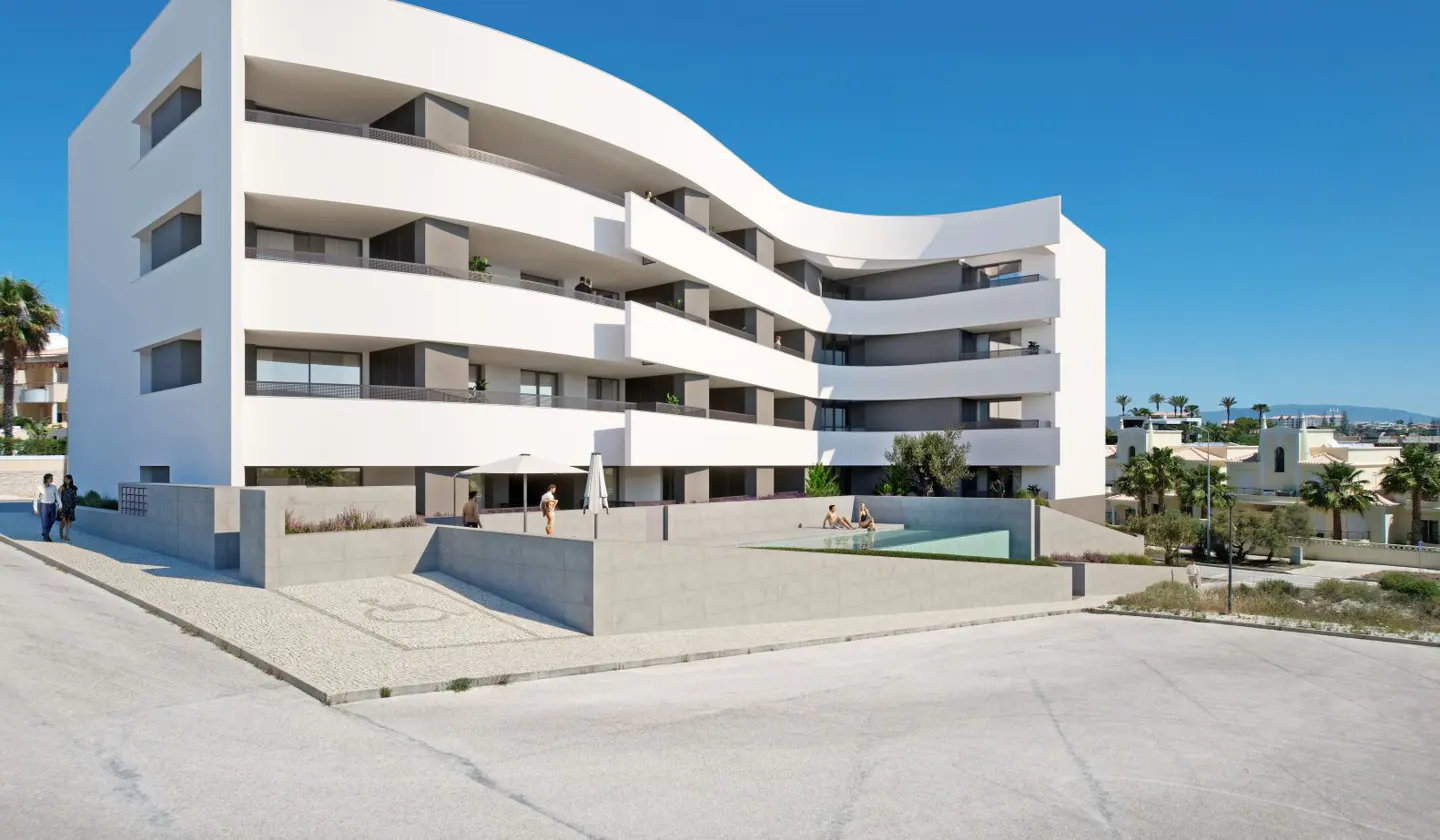 Modern white apartment building with curved balconies, a ground-level swimming pool, and a paved parking area.