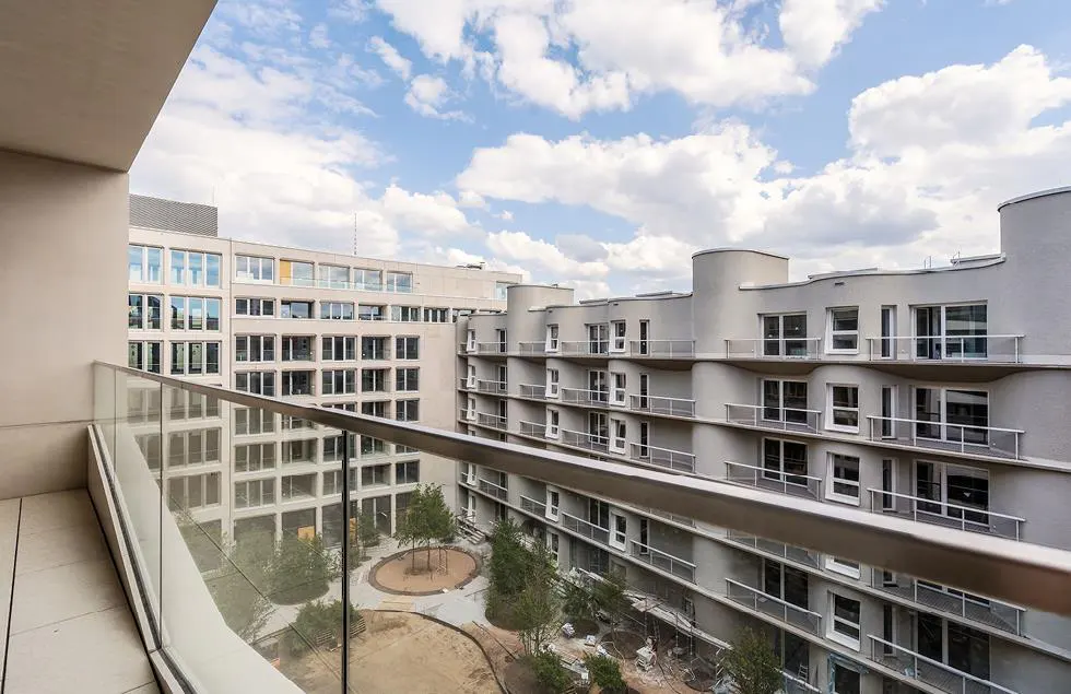View from a balcony with glass railings overlooking a courtyard surrounded by modern apartment buildings under a cloudy sky.