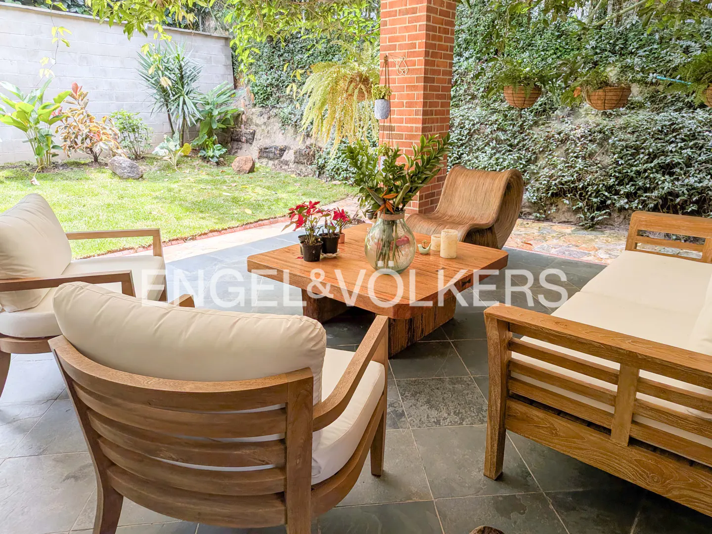 Outdoor patio with wooden furniture, including chairs, a sofa, and a coffee table with flowers and candles. Lush greenery in the background.