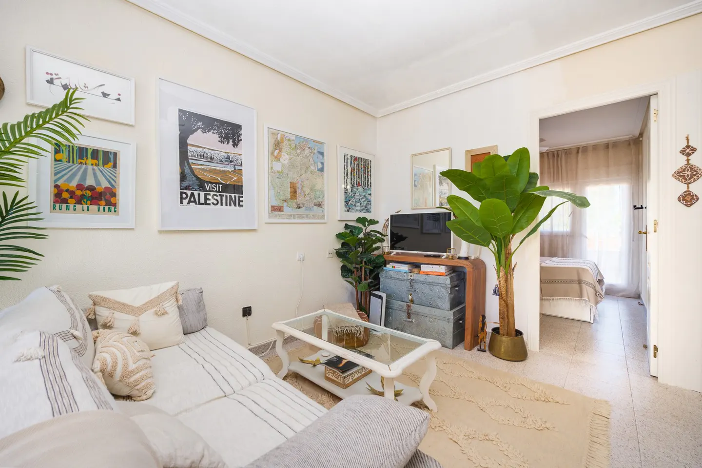 Living room with a white sofa, glass coffee table, and a TV on a wooden stand with metal trunks. Art on the wall and a view into a bedroom.