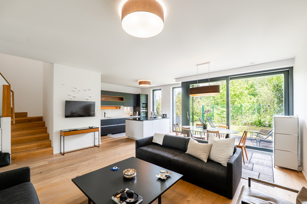 Bright, modern open-plan living space with wood floors, black sofa, kitchen, and sliding glass doors to a green garden.