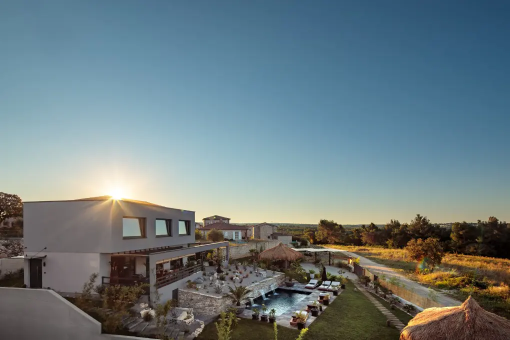 Modern two-story house with a pool, waterfall feature, and thatched-roof cabana under a clear blue sky at sunset.